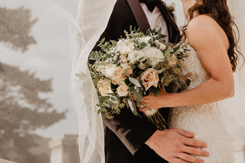 Close-up of bride and groom on their wedding day, with the bride holding a bouquet of white and blush pink roses and greenery, while the groom embraces her under a soft, flowing veil.